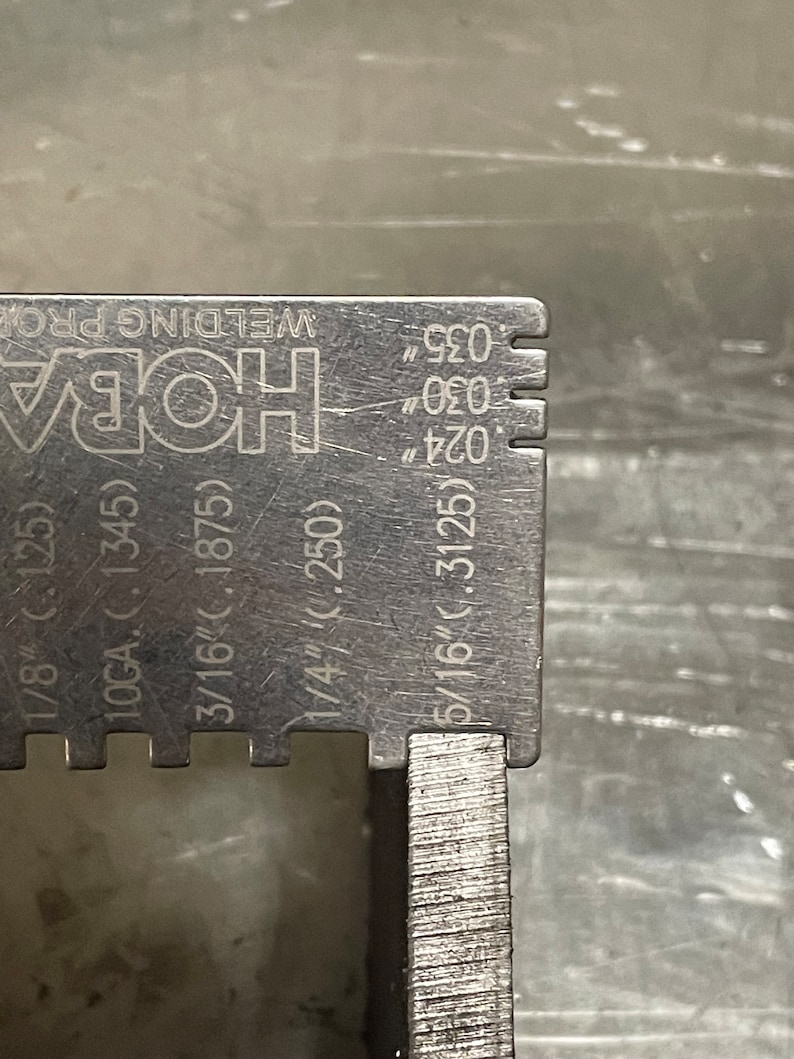 May include: A close-up of a stainless steel welding gauge with measurements in inches. The gauge has markings for 1/8", 3/16", 1/4", and 5/16". The text "HOBA WELDING PRO" is visible.