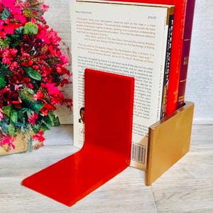 May include: A red acrylic bookend with a gold-colored base supports several books. The books have red and white spines. The bookend is on a light-colored surface, with a bouquet of pink flowers in the background.