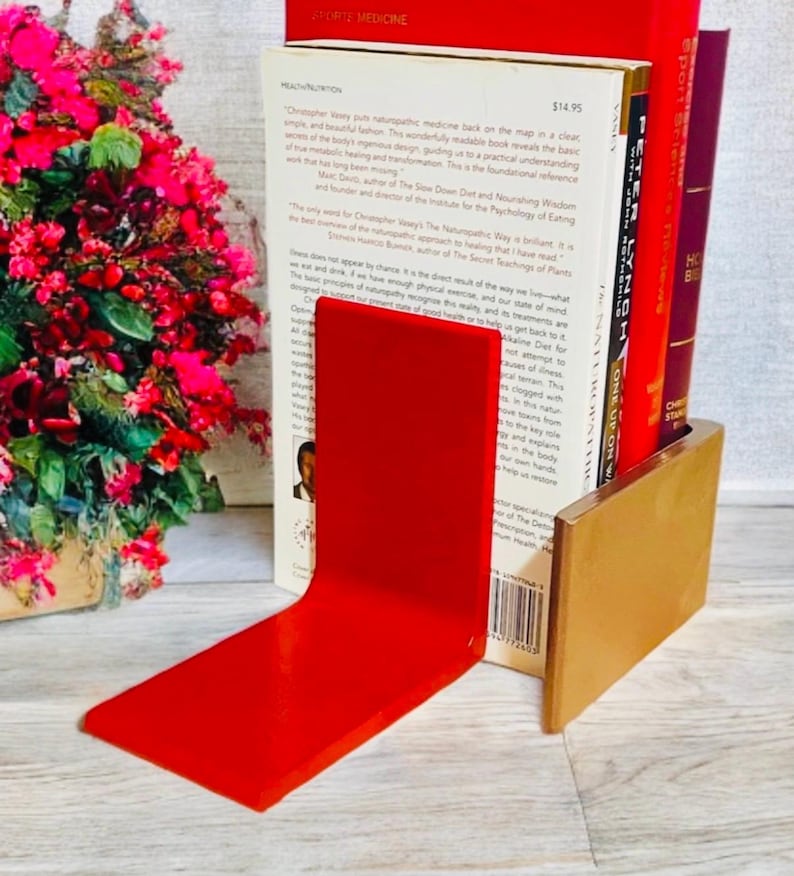 May include: A red acrylic bookend with a gold-colored base supports several books. The books have red and white spines. The bookend is on a light-colored surface, with a bouquet of pink flowers in the background.