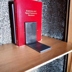 May include: A red book titled "Exercise and Sport Sciences Reviews" is held in place by a dark gray metal bookend. The bookend is L-shaped and sits on a light brown wooden shelf. The background is a textured gray surface.