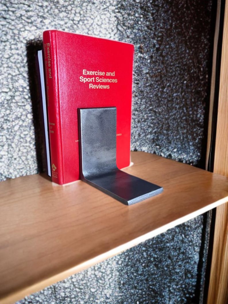 May include: A red book titled "Exercise and Sport Sciences Reviews" is held in place by a dark gray metal bookend. The bookend is L-shaped and sits on a light brown wooden shelf. The background is a textured gray surface.