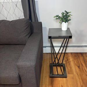 May include: A gray couch with a side table made of metal and a gray top. The table has a plant in a white pot on top of it.