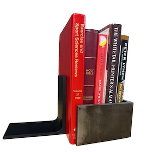 May include: A black L-shaped metal bookend supporting a row of five books. The books include Exercise and Sport Sciences Reviews Volume 27 1999, a Holy Bible, Sonia Taitz's Mother's Daughter, The Whitetail Hunter's Almanac, and Peter Lynch's One Up on Wall Street.