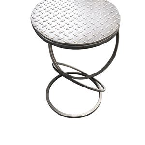 May include: A small, industrial-style metal stool with a circular, diamond-plate seat. The seat is supported by a looping, curved metal base. The stool has a minimalist design and a silver finish, suitable for various interior settings.