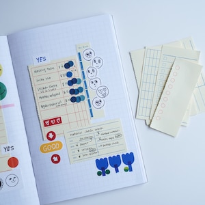 May include: An open notebook with a grid pattern on the pages. The notebook has various stickers and notes on the pages, including a checklist, a table with prices, and a list of materials. The notebook is on a white surface.