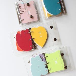 Può includere: Una collezione di piccoli raccoglitori in plastica trasparente, ciascuno contenente forme di carta fustellate colorate. Le forme includono orsi, cuori e disegni floreali in vari colori, come rosa, giallo e blu. I raccoglitori sono tenuti insieme da anelli metallici.