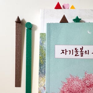 Puede incluir: Dos marcapáginas largos, delgados y texturizados, uno marrón con punta cónica y otro verde con punta circular, junto a un libro. El libro tiene una cubierta azul claro con flores rosas y texto en coreano, con pequeños marcadores geométricos visibles.