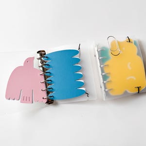 May include: Three small notebooks with die-cut pages in pink, blue, and yellow. The first has a bird shape, the second has wavy pages, and the third a blob shape. They are bound with silver rings and clear plastic covers. The notebooks are ideal for jotting down notes.