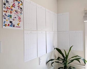 May include: A white wall with nine blank calendars hanging on it. The calendars are numbered 01 through 09. A green plant is in the corner of the room.
