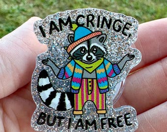 May include: A glittery badge reel featuring a cartoon raccoon wearing a hat, scarf, and sweater. The text on the badge reads "I AM CRINGE, BUT I AM FREE". The badge has a clear plastic reel and a metal clip.