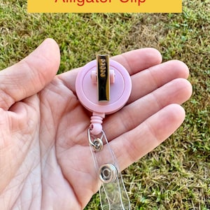 May include: A pink alligator clip badge reel with a clear plastic strap and metal hardware. The text "Alligator Clip" is in a yellow rectangle. The reel has a gold-colored metal clip.