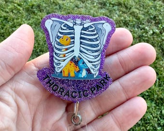 May include: A purple glitter badge reel featuring a rib cage design with three cartoon dinosaurs inside. The text "THORACIC PARK" is written in purple letters. The badge reel has a clear plastic strap and a metal clip.