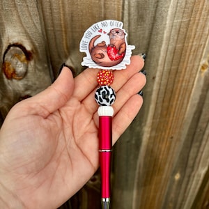 May include: A red metal pen featuring a decorative topper with a cartoon otter holding a red heart and the text LOVE YOU LIKE NO OTTER. The pen barrel is accented with three beads: a red glitter bead, a black and white cow print bead, and a white bead.