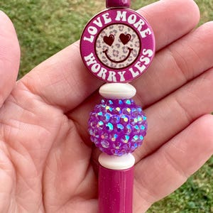 May include: A magenta pen with a decorative top. The top features a round design with the words "Love More Worry Less" around a smiley face with heart eyes. Below the design is a white bead, a purple crystal ball, and a magenta pen body.