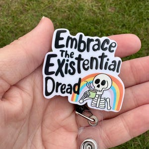 May include: A white badge reel with the text "Embrace The Existential Dread." The badge features a cartoon skeleton drinking from a mug, with a rainbow in the background. The reel has a clear plastic strap and a metal clip.