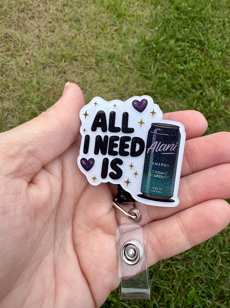 May include: A white badge reel with the text "ALL I NEED IS" and two purple heart accents. The badge reel also features a small can of "Alani Energy Cosmic Stardust" and a clear plastic strap.
