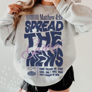 May include: A gray sweatshirt with the text "Spread the Good News" in blue and purple lettering. The text "Matthew 4:19" is also printed on the sweatshirt. The sweatshirt is being worn by a person.