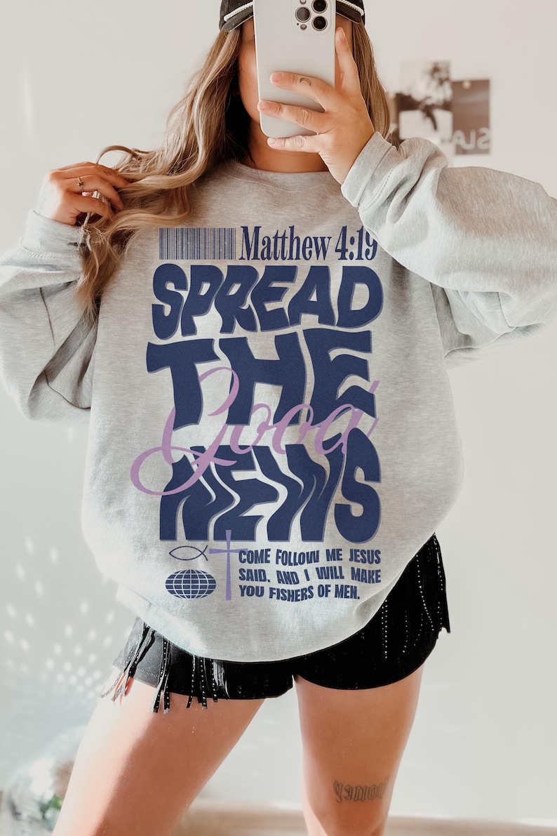 May include: A gray sweatshirt with the text "Spread the Good News" in blue and purple lettering. The text "Matthew 4:19" is also printed on the sweatshirt. The sweatshirt is being worn by a person.