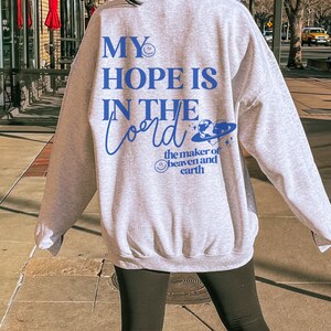 May include: A pale grey sweatshirt featuring the blue text "MY HOPE IS IN THE Lord". The design includes a smiley face, a planet, and the words "the maker of heaven and earth". The sweatshirt is a relaxed fit.