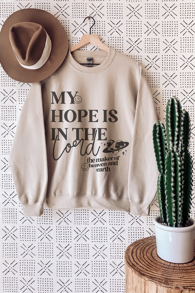 May include: A beige sweatshirt with the text "MY HOPE IS IN THE Lord the maker of heaven and earth" in black script. A brown hat hangs to the left. A cactus in a white pot sits to the right. The background is a white and black patterned wall.