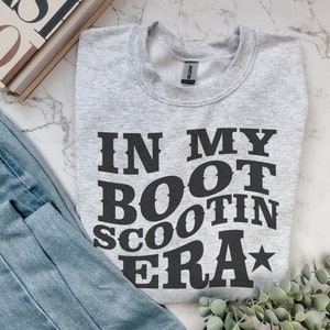 May include: A gray sweatshirt with the text "IN MY BOOT SCOOTIN ERA" printed in black. The text is in a bold, western-style font with a star next to the word "ERA".