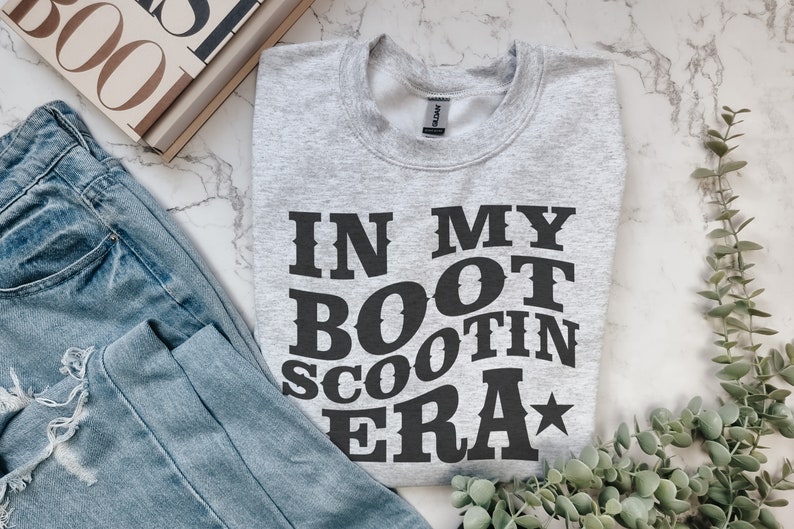 May include: A gray sweatshirt with the text "IN MY BOOT SCOOTIN ERA" printed in black. The text is in a bold, western-style font with a star next to the word "ERA".