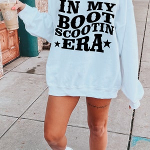 May include: A white sweatshirt with the text "IN MY BOOT SCOOTIN ERA" printed in black. The sweatshirt has a relaxed fit and is worn by a person with a tattoo on their leg.