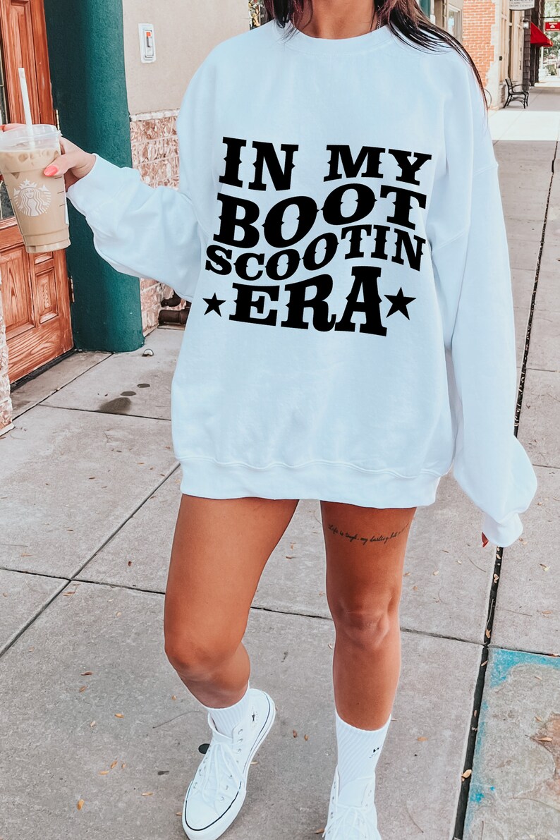May include: A white sweatshirt with the text "IN MY BOOT SCOOTIN ERA" printed in black. The sweatshirt has a relaxed fit and is worn by a person with a tattoo on their leg.