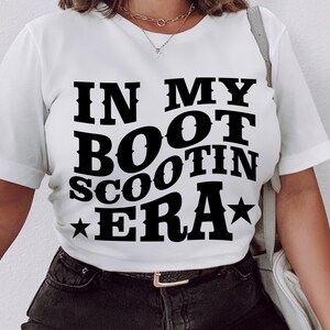 May include: White t-shirt with black text that reads "In My Boot Scootin Era" with a star on either side of the text.