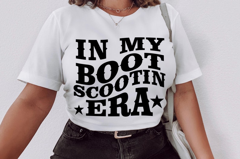 May include: White t-shirt with black text that reads "In My Boot Scootin Era" with a star on either side of the text.