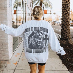 May include: Light gray sweatshirt with a large graphic on the back featuring a lion, cross, and the text "GOD IS OUR REFUGE AND STRENGTH". The sweatshirt also has additional text from Psalm 46:1-3.