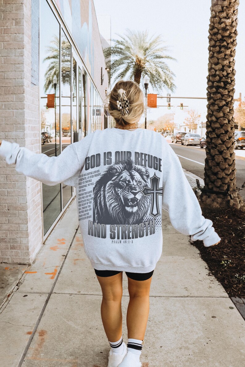 May include: Light gray sweatshirt with a large graphic on the back featuring a lion, cross, and the text "GOD IS OUR REFUGE AND STRENGTH". The sweatshirt also has additional text from Psalm 46:1-3.