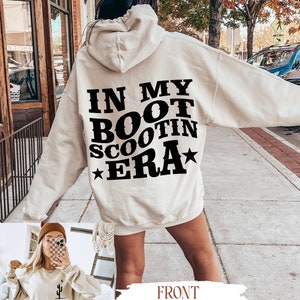 May include: A cream-colored hoodie with a black graphic that says "In My Boot Scootin Era" with a star on either side of the text. The hoodie has a front pocket and a drawstring hood. The model is wearing the hoodie with a pair of white sneakers.