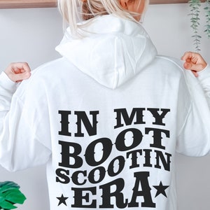 May include: White hoodie with black text that reads "In My Boot Scootin Era" with a star on either side of the text.