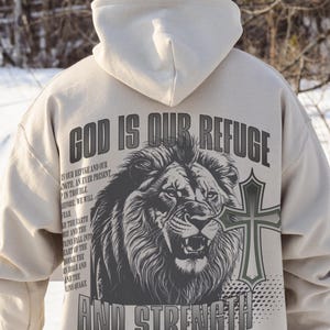 May include: Beige hoodie with the text "GOD IS OUR REFUGE AND STRENGTH" and a lion graphic. The back of the hoodie also features a cross and the Bible verse Psalm 46:1-3. The hoodie has a drawstring hood and long sleeves.