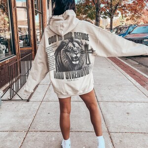 May include: Cream-colored hoodie with a large graphic on the back featuring a lion, cross, and text that reads "GOD IS OUR REFUGE AND STRENGTH". The hoodie has a hood and long sleeves. The person is wearing white sneakers.
