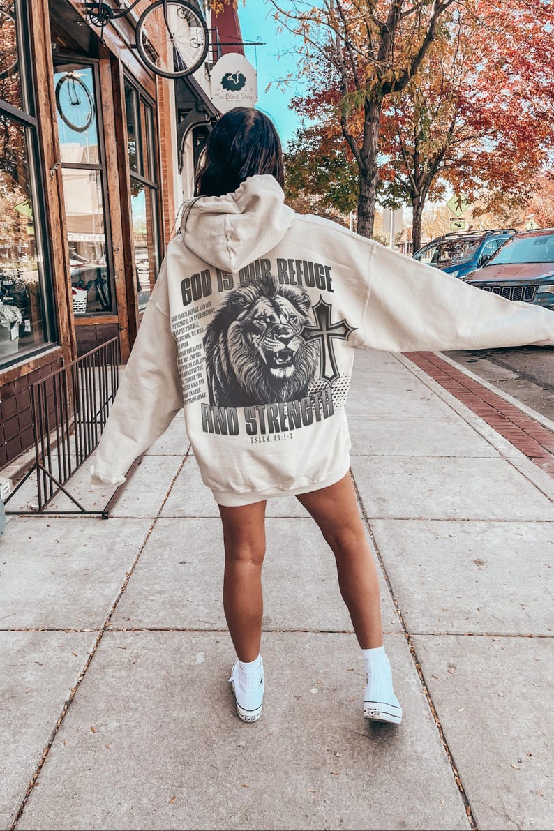 May include: Cream-colored hoodie with a large graphic on the back featuring a lion, cross, and text that reads "GOD IS OUR REFUGE AND STRENGTH". The hoodie has a hood and long sleeves. The person is wearing white sneakers.