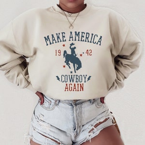 May include: A beige sweatshirt with the text "MAKE AMERICA COWBOY AGAIN" in red and blue lettering. The sweatshirt features a silhouette of a cowboy riding a bucking horse with stars and the numbers 19 and 42.