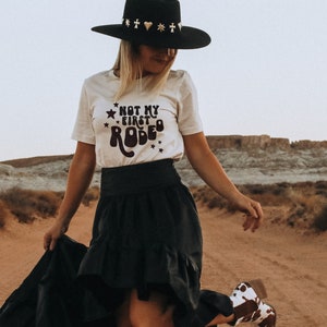 K&ouml;nnte beinhalten: Eine Frau tr&auml;gt ein wei&szlig;es T-Shirt mit dem Text "Not my first rodeo" in schwarzen Buchstaben und einen schwarzen Hut mit goldenen und silbernen Verzierungen. Sie tr&auml;gt einen schwarzen Rock und Cowboystiefel. Die Frau steht in einer W&uuml;stenlandschaft.