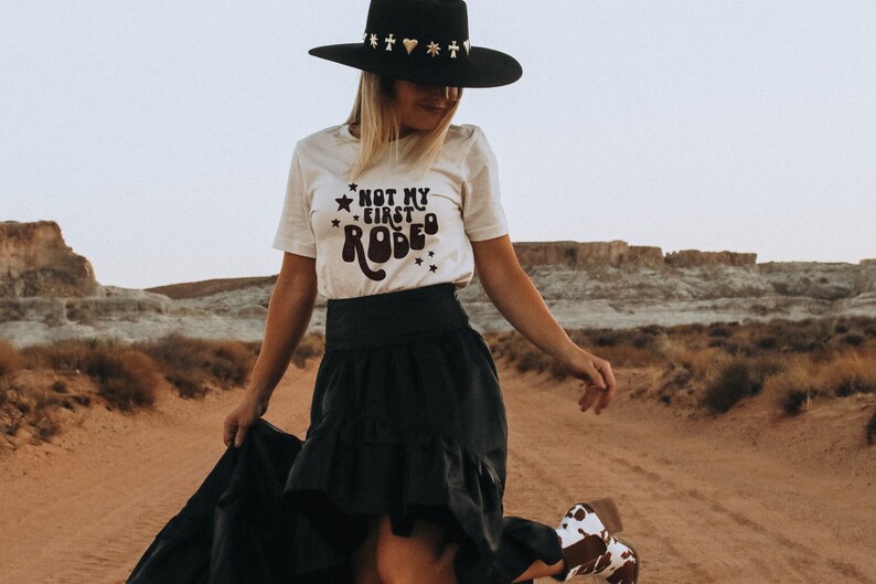 K&ouml;nnte beinhalten: Eine Frau tr&auml;gt ein wei&szlig;es T-Shirt mit dem Text "Not my first rodeo" in schwarzen Buchstaben und einen schwarzen Hut mit goldenen und silbernen Verzierungen. Sie tr&auml;gt einen schwarzen Rock und Cowboystiefel. Die Frau steht in einer W&uuml;stenlandschaft.