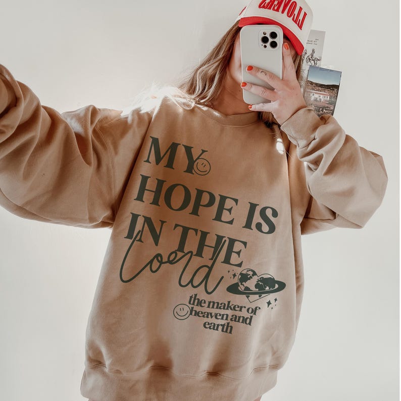 May include: A beige sweatshirt featuring the words "MY HOPE IS IN THE Lord" in dark green. The design includes a smiley face, a heart-shaped planet, and the words "the maker of heaven and earth". A red and white hat is visible.