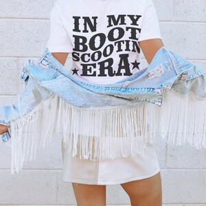 May include: A white t-shirt with the text "IN MY BOOT SCOOTIN' ERA" in black lettering. The person is wearing a white cowboy hat and a light blue denim jacket with white fringe.