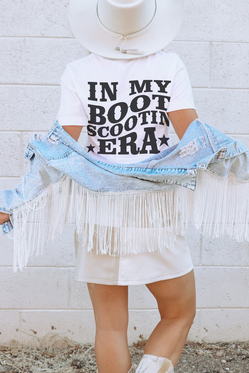 May include: A white t-shirt with the text "IN MY BOOT SCOOTIN' ERA" in black lettering. The person is wearing a white cowboy hat and a light blue denim jacket with white fringe.