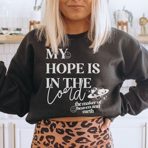 May include: Dark grey sweatshirt featuring the white text "MY HOPE IS IN THE Lord". The design includes a smiling face and a graphic of the earth and a planet. The sweatshirt is worn with leopard print leggings.