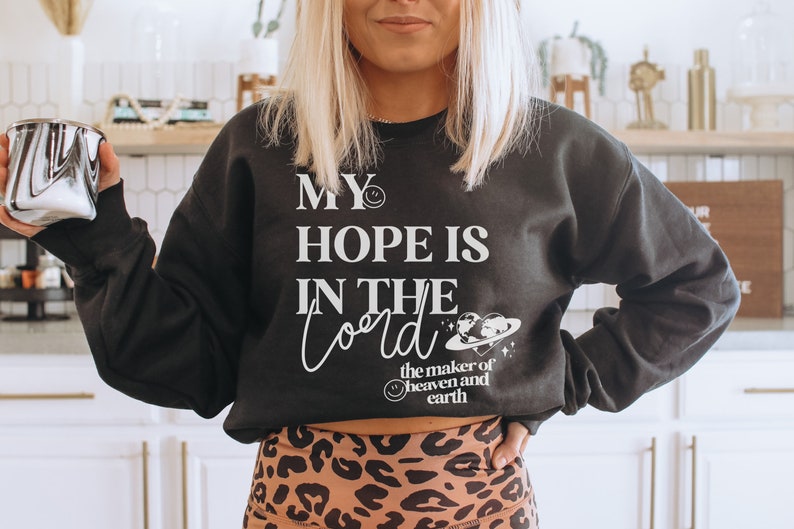 May include: Dark grey sweatshirt featuring the white text "MY HOPE IS IN THE Lord". The design includes a smiling face and a graphic of the earth and a planet. The sweatshirt is worn with leopard print leggings.
