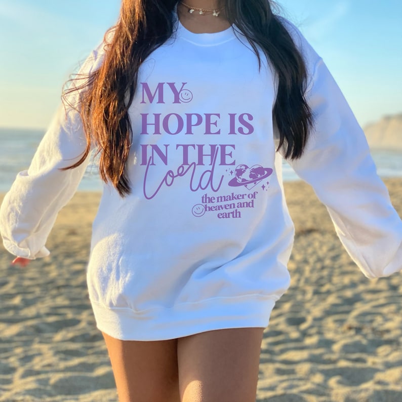 May include: White sweatshirt featuring the purple text "MY HOPE IS IN THE Lord". The design includes a smiling face, a planet with a heart, and the words "the maker of heaven and earth".