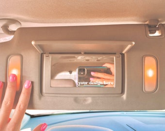 May include: A beige car sun visor with a mirror and two lights. The mirror is open and shows a reflection of a hand holding a mobile phone with the text "your design here" on the screen.