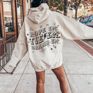 May include: A cream colored hoodie with a black graphic print that says "Rope 'em, Tie 'em, Brand 'em".