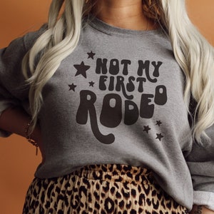 K&ouml;nnte beinhalten: Ein graues Sweatshirt mit dem Text "Not My First Rodeo" in einer Retro-Schriftart mit Sternen.