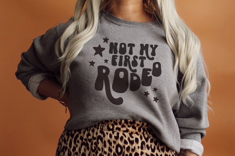 K&ouml;nnte beinhalten: Ein graues Sweatshirt mit dem Text "Not My First Rodeo" in einer Retro-Schriftart mit Sternen.